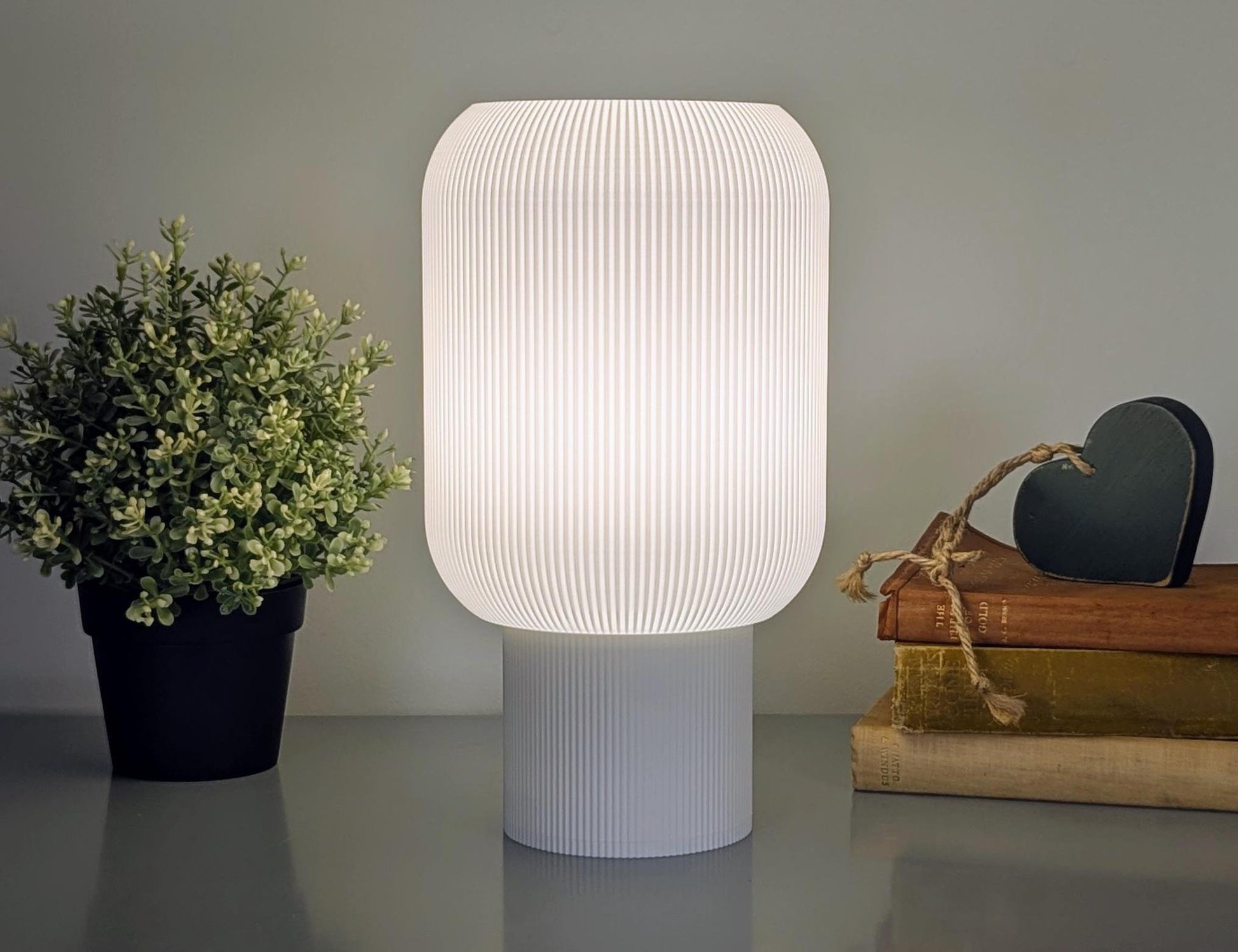 A white Cumulis Table Lamp from Dryft Home, featuring 3D printed pleated design with sustainable materials, casts a soft glow beside a potted plant and heart-shaped decor on stacked books, creating a cozy minimalist vibe.