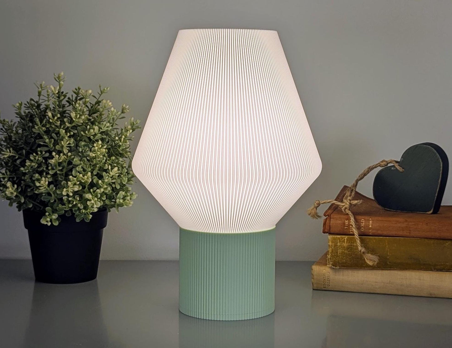 The Inversis Table Lamp from Dryft Home features a geometric 3D printed shade and emits soft white light. Resting on its teal base alongside a potted plant and twine-wrapped books, it is complemented by a small green heart decor, enhancing the charm of this sustainable setup.