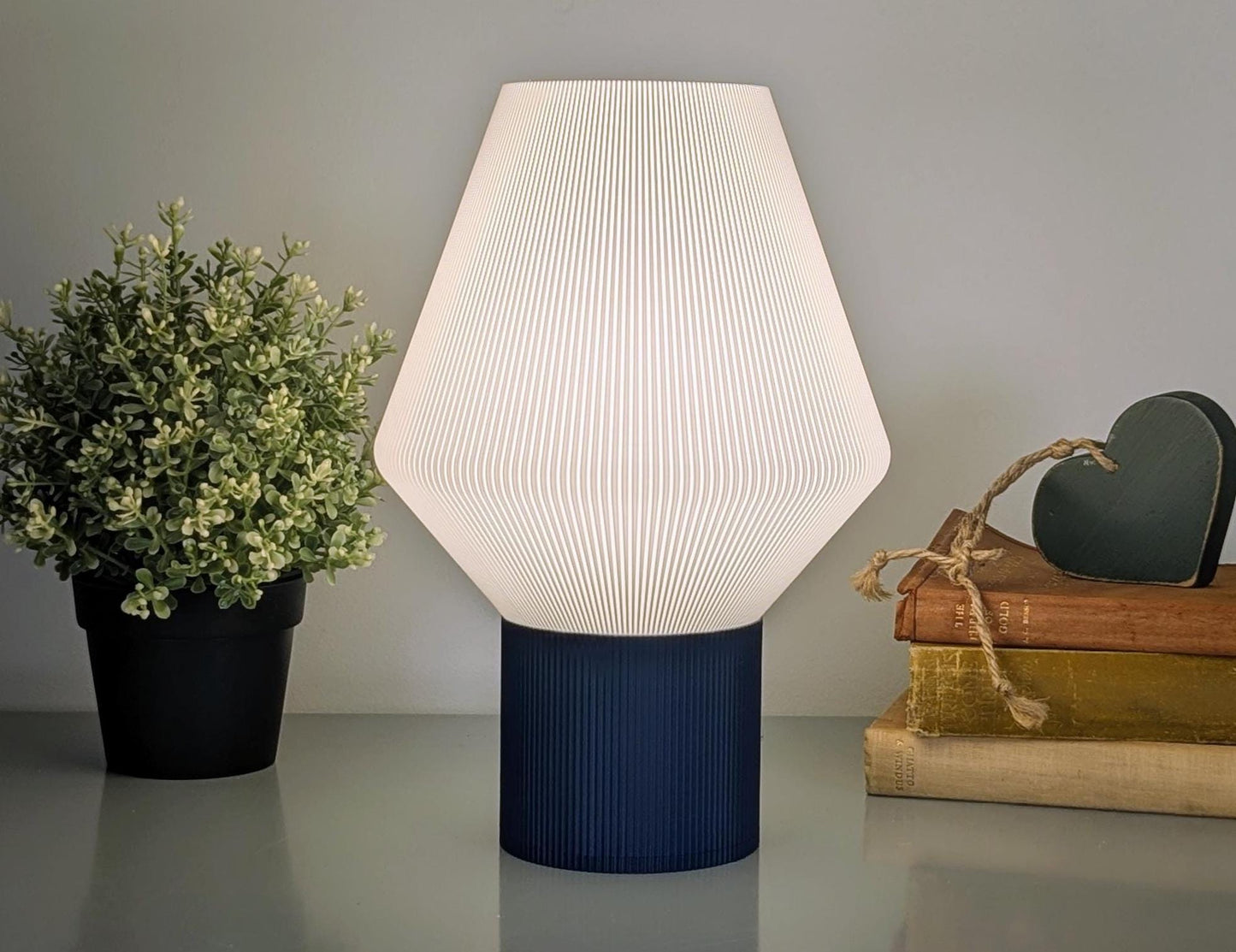 The Dryft Home Inversis Table Lamp features a geometric, ribbed white shade and dark base on a reflective surface, offering sustainable elegance. Lit with a warm glow, its paired with a potted plant and twine-bound books topped with a heart-shaped ornament for added charm.