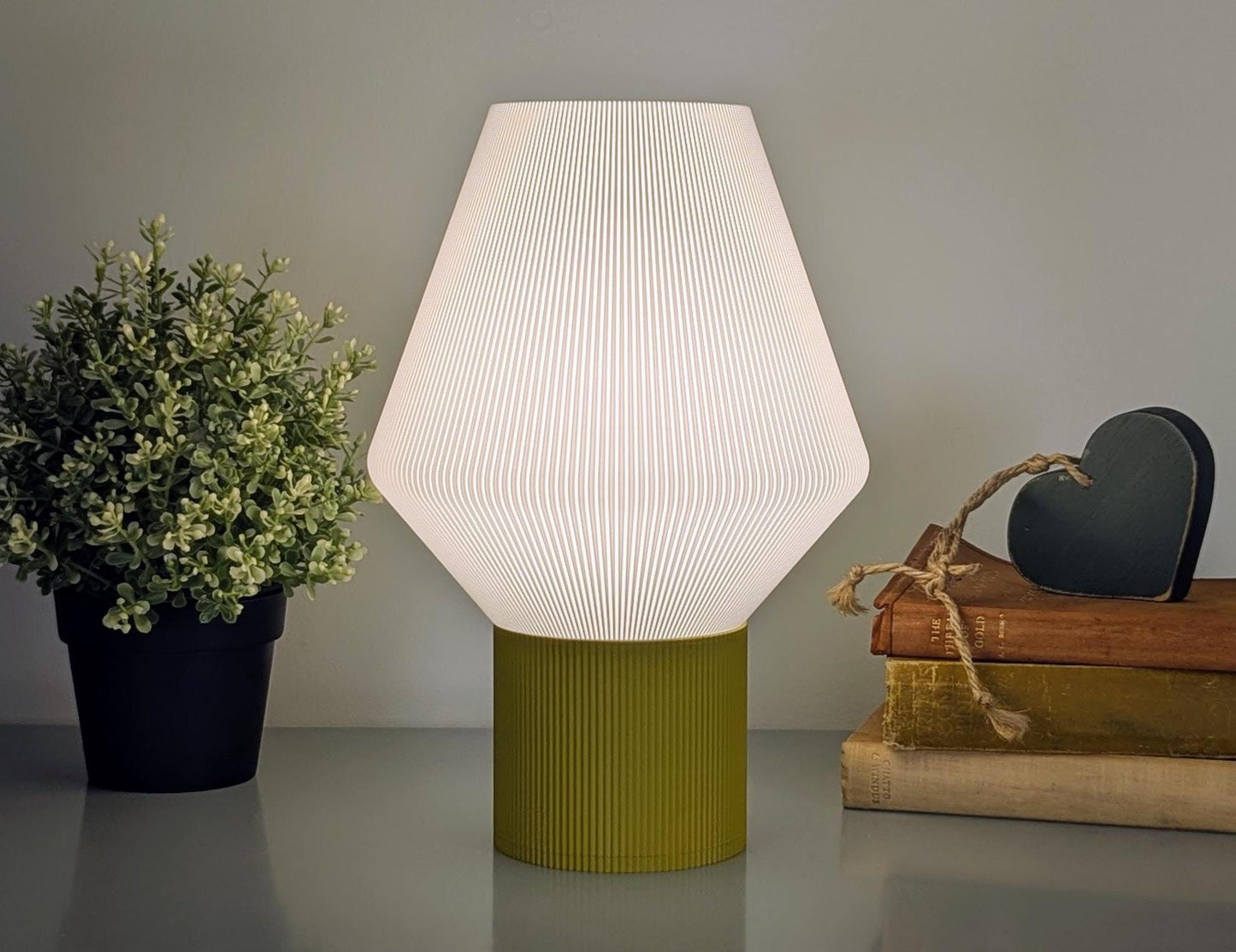 The Inversis Table Lamp by Dryft Home, featuring a 3D printed geometric white shade and green base, casts a warm glow. Beside it sits a small potted plant, while stacked books with a wooden heart decoration enhance the ambiance of sustainable decor.